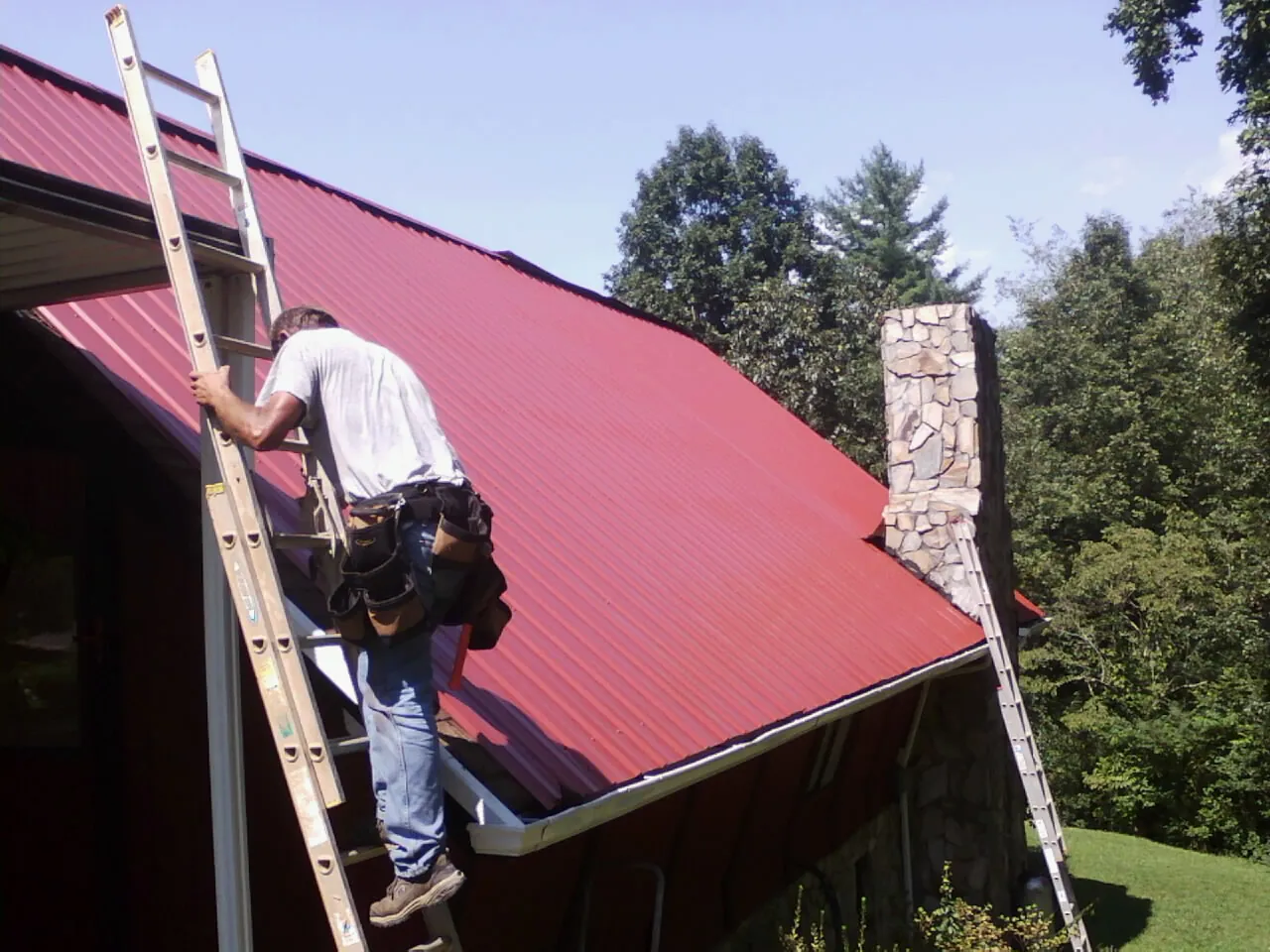 Roofer on a ladder beside a red metal roof and stone chimney — Soffit & Fascia Repair by Roofing Brothers in Clyde Park